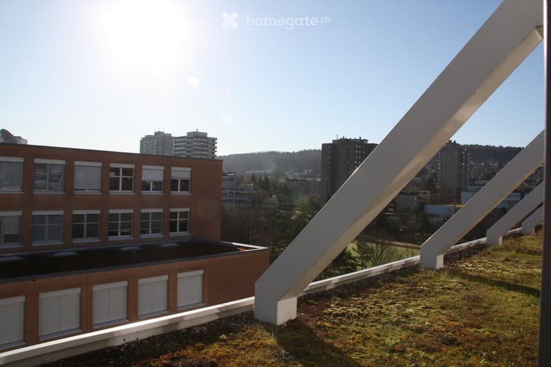A modern building with a green roof and large white geometric structures extending from the roof, surrounded by other buildings and a clear blue sky.