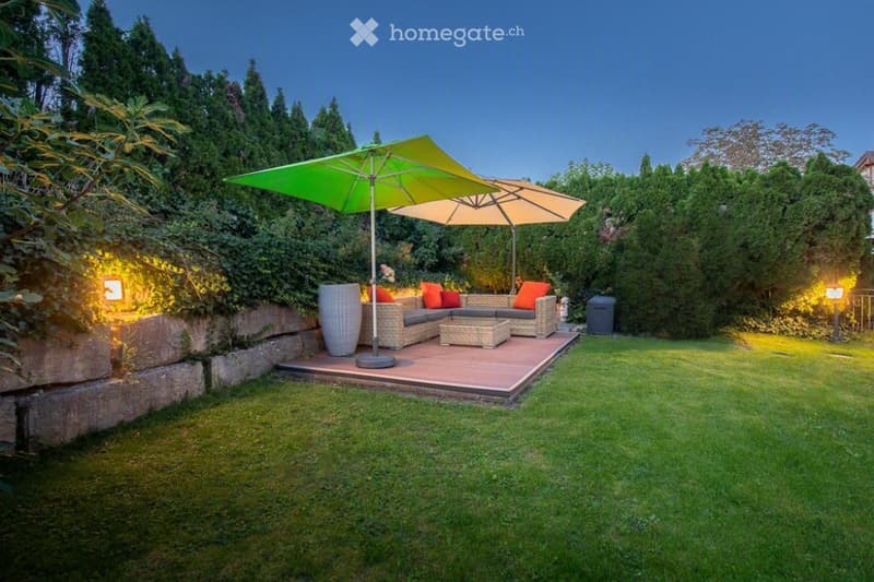 Well-maintained garden with a raised wooden deck, outdoor sofa set, coffee table, large umbrella, and decorative vase.