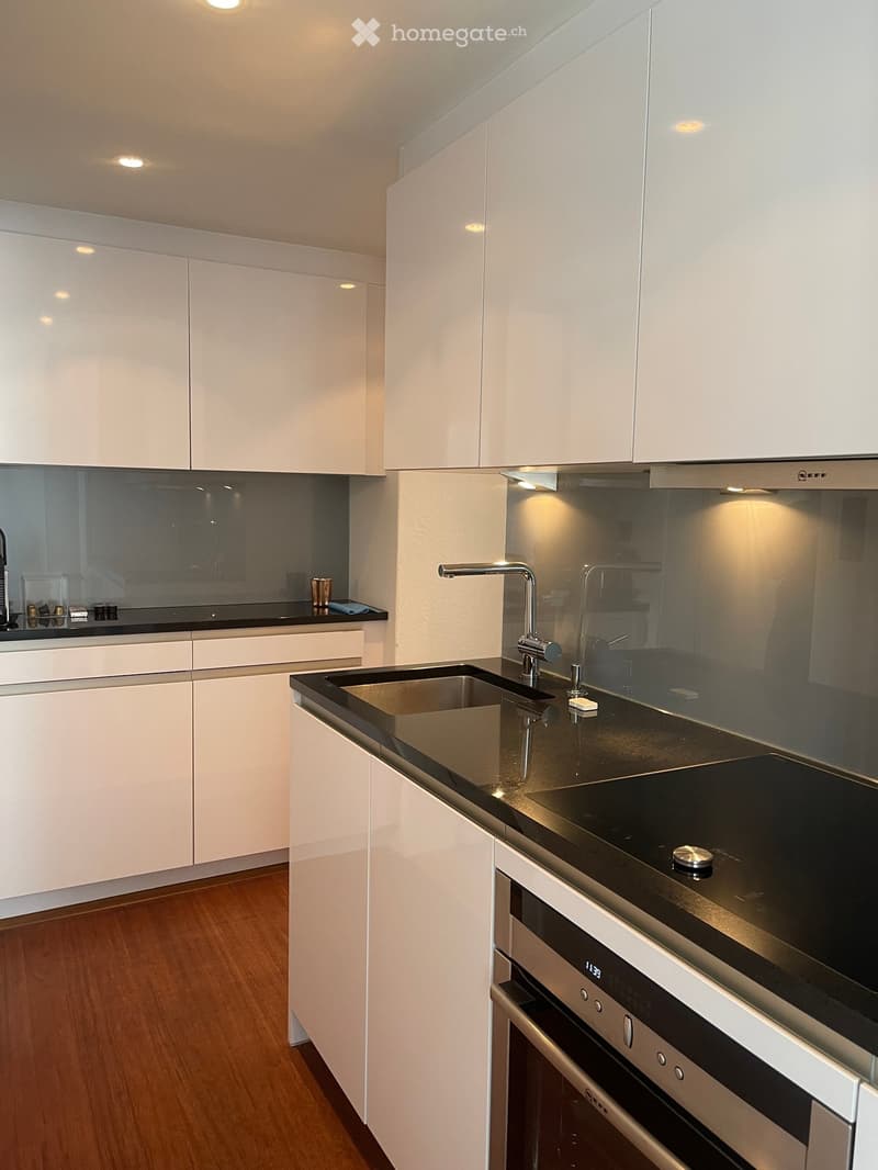 Modern kitchen with white cabinets, black countertop, stainless steel sink and faucet, built-in oven, and wooden flooring.