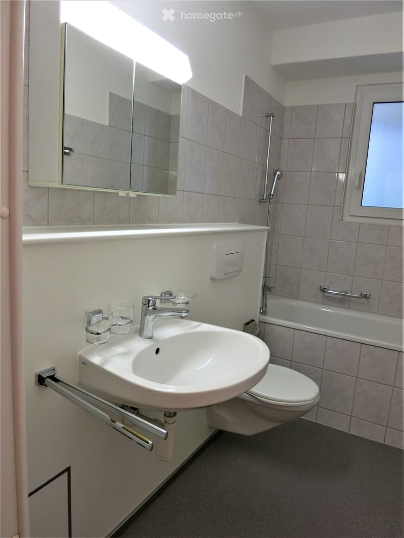 Modern bathroom with a white sink, chrome faucet, mirror, toilet, bathtub, and a small window. Includes a handrail and a cup on the sink.
