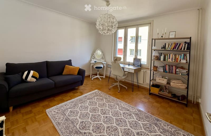 A modern living room with a dark blue sofa, patterned cushions, a large bookshelf filled with books, a desk with a laptop, a white chair, and a decorative mirror. The room features wooden flooring and large windows with a view of the street.