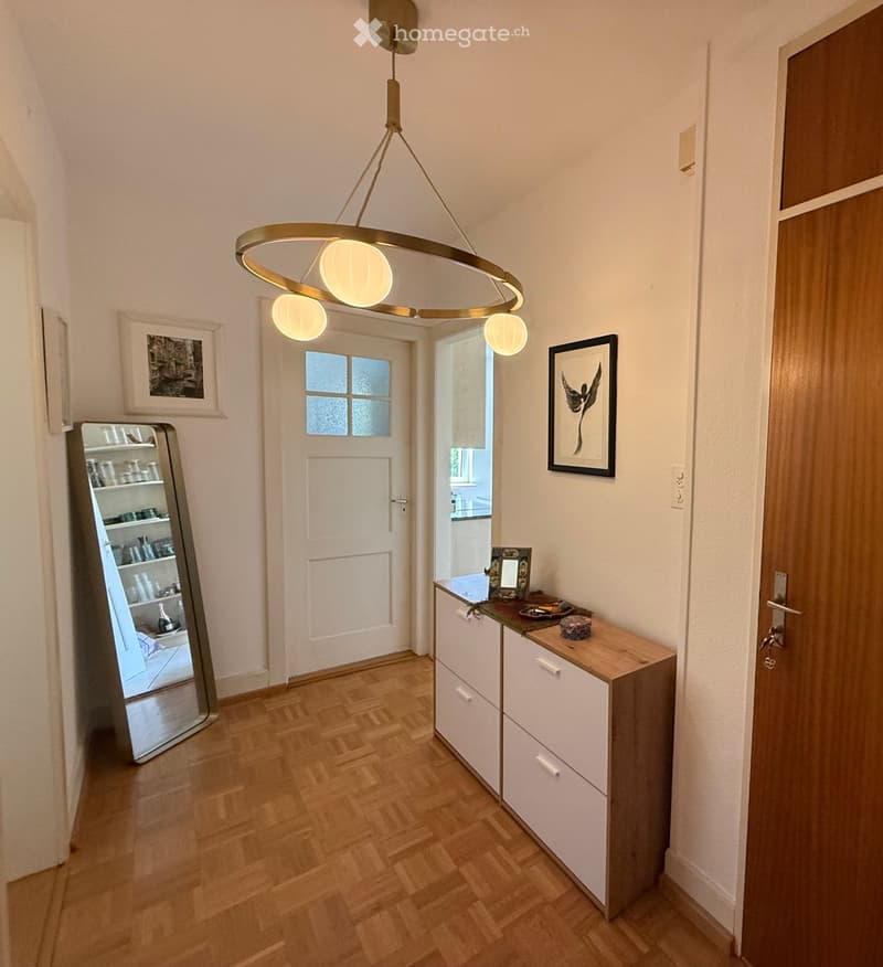 Modern hallway with wooden flooring, white walls, a large round pendant light fixture, a small console table with drawers, and framed artwork on the walls. A large mirror reflects part of the adjacent room.
