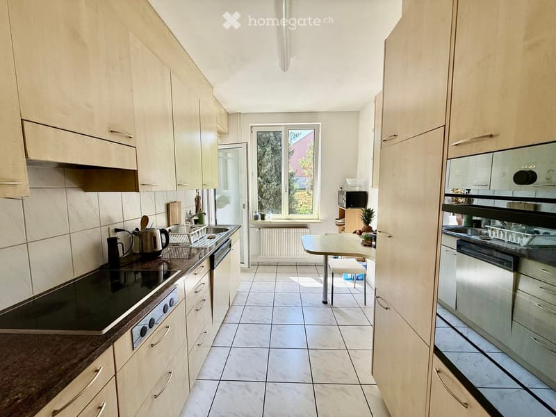 Modern kitchen with light wood cabinets, tiled floor, and stainless steel appliances. Features an island with a sink, stove, and microwave, along with a small dining area with a table and chair.