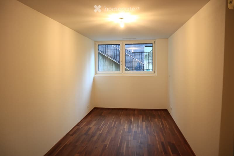 Empty room, hardwood floors, white walls, natural light from window, light fixture on ceiling.