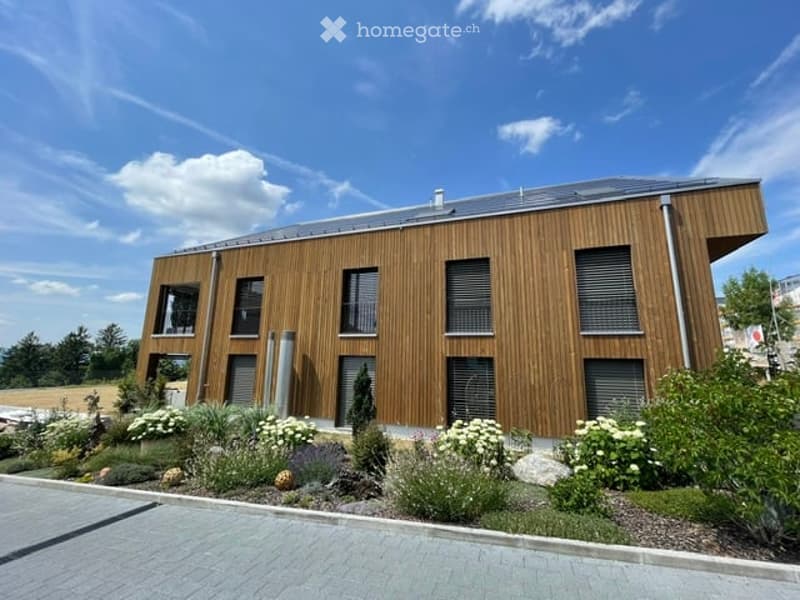 Modern wooden building with a flat roof, large windows, and a landscaped garden area with white flowers and shrubs.