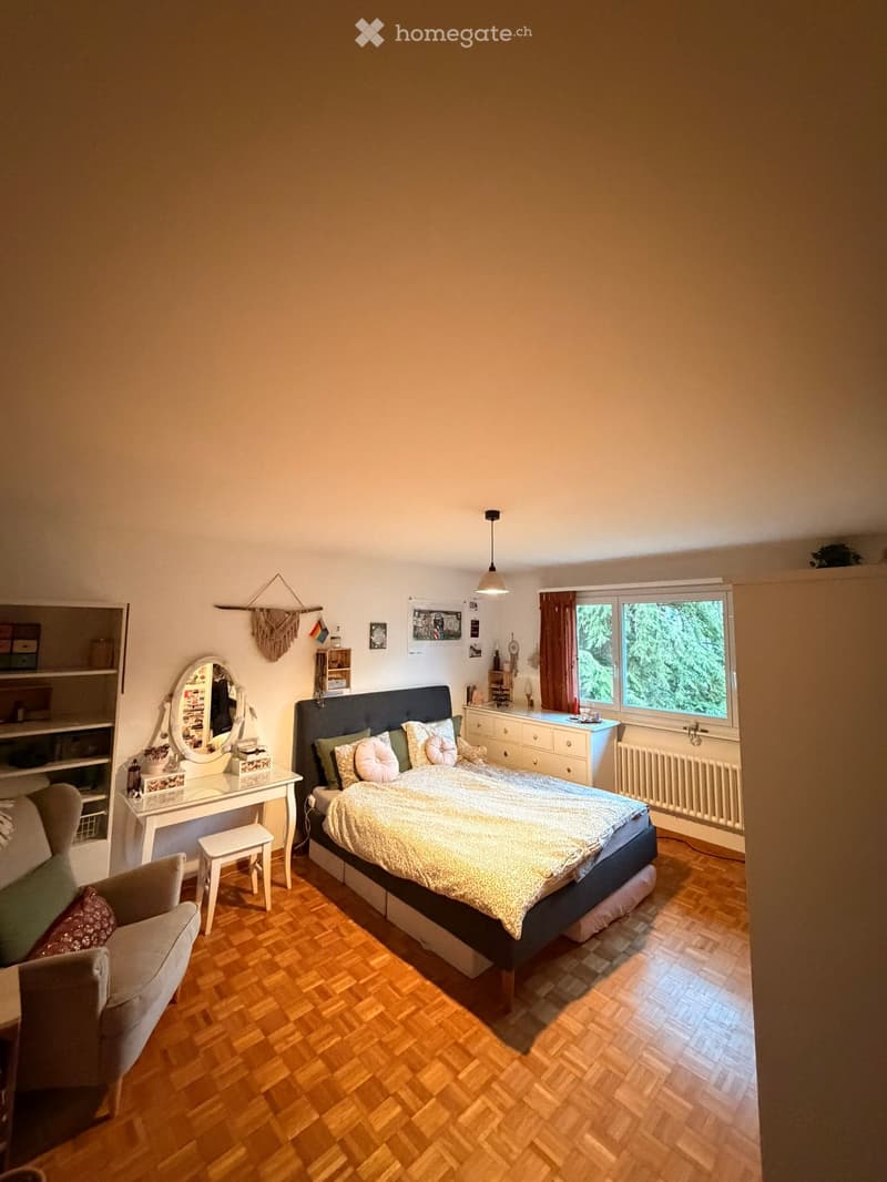 Cozy bedroom with wooden flooring, a large bed with pillows and a patterned bedspread, a vanity table with a mirror, a small chair, and a window with curtains.