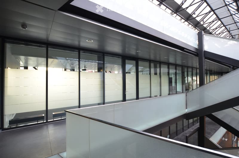 Modern interior corridor with glass walls, sleek metal railings, and a spacious open design. The area features a raised ceiling with integrated lighting and a polished concrete floor.