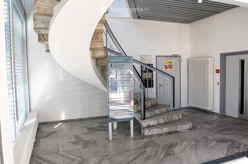 Modern hallway with a curved staircase, glass railing, and polished stone tiles. Features a signboard and a radiator on the wall.