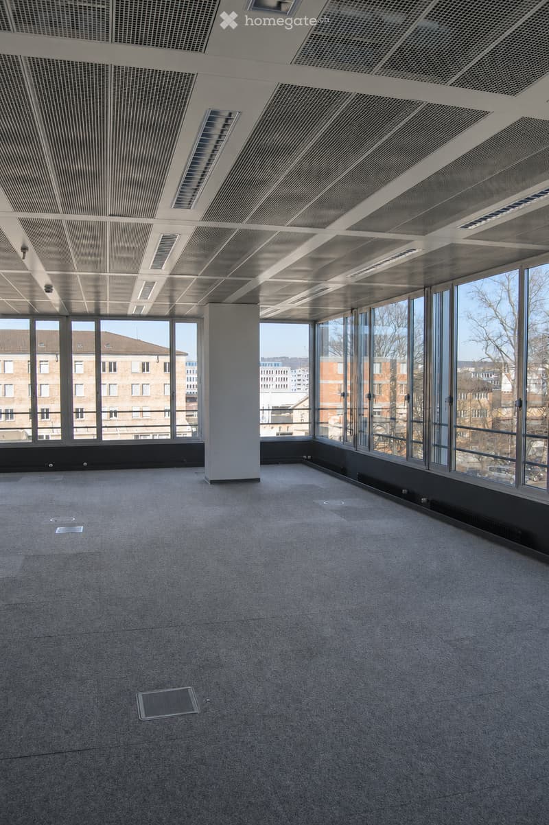 Empty room with grey carpeted floor, large windows, and a central column. No furniture or equipment present.