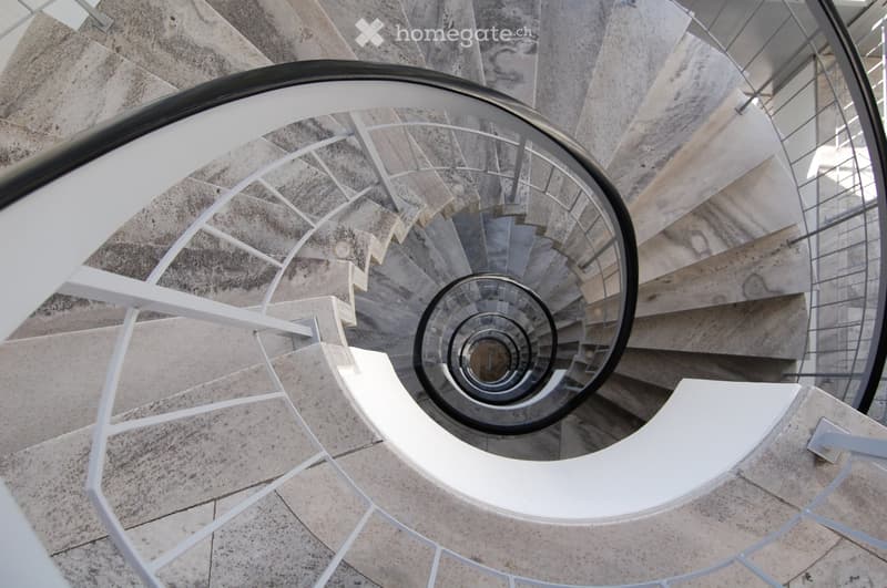 Spiral staircase with modern white railings and stone steps, leading upwards with a glass ceiling allowing natural light.