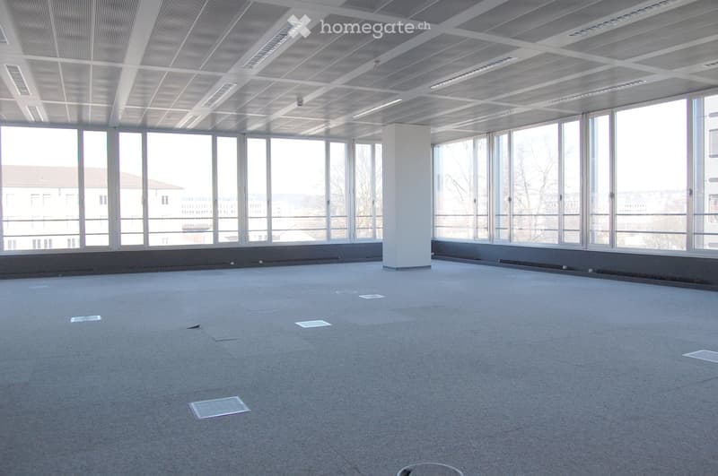 Empty room with grey carpeted floor, large windows, white ceiling with recessed lighting, and a central white pillar.