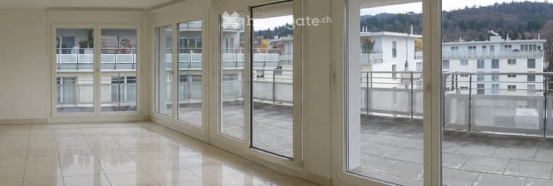 Bright, empty room with large glass doors leading to a tiled balcony, offering a scenic view of surrounding buildings and greenery.