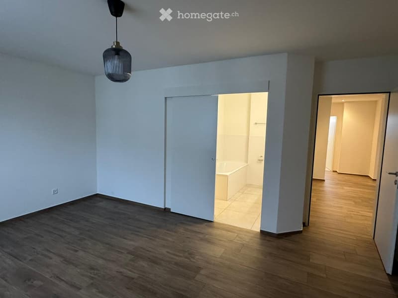 Empty hallway with wooden flooring, light blue walls, and a hanging pendant light. Open doors lead to a tiled bathroom and another room.