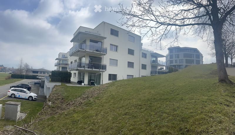 White multi-story building with balconies, located on the side of the hill, with a parking area below