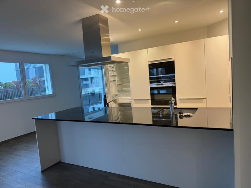 Modern kitchen with white cabinets, a central island, built-in oven, and a range hood. The space features large windows with a view of the balcony and surrounding buildings.