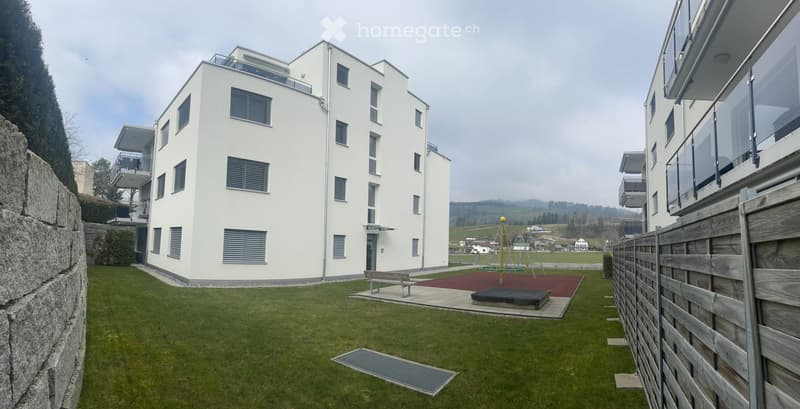 3 story white building with balconies and entrance area, bench, red flooring in front of building, large lawn and playground in front, tall stone wall on the left, wooden fence on the right
