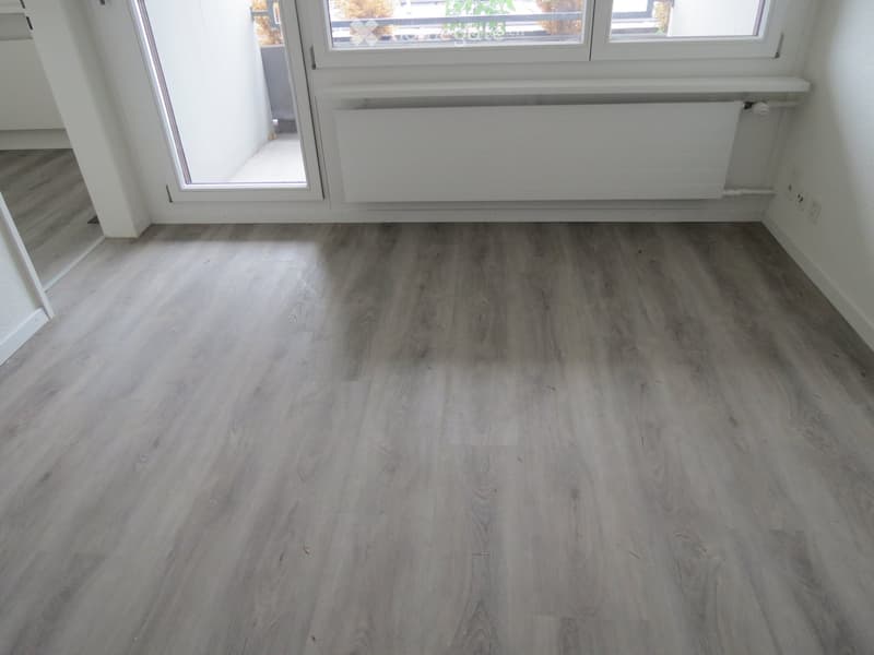 Empty room with light grey wooden flooring, white walls, large windows, and a radiator. No furniture or equipment present.