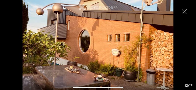 Outdoor patio area with a modern brick house in the background, featuring a large round window, potted plants, a stack of firewood, a tall outdoor lamp, and a small table with a few items on it.