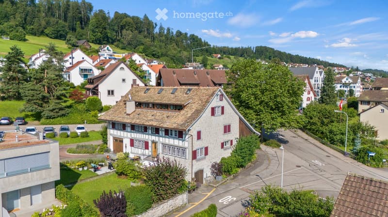 Aerial view of a residential area with multiple houses, surrounded by greenery and hills. The houses have varied architectural styles, with some featuring red shutters and tiled roofs. There is a parking area visible in the foreground.