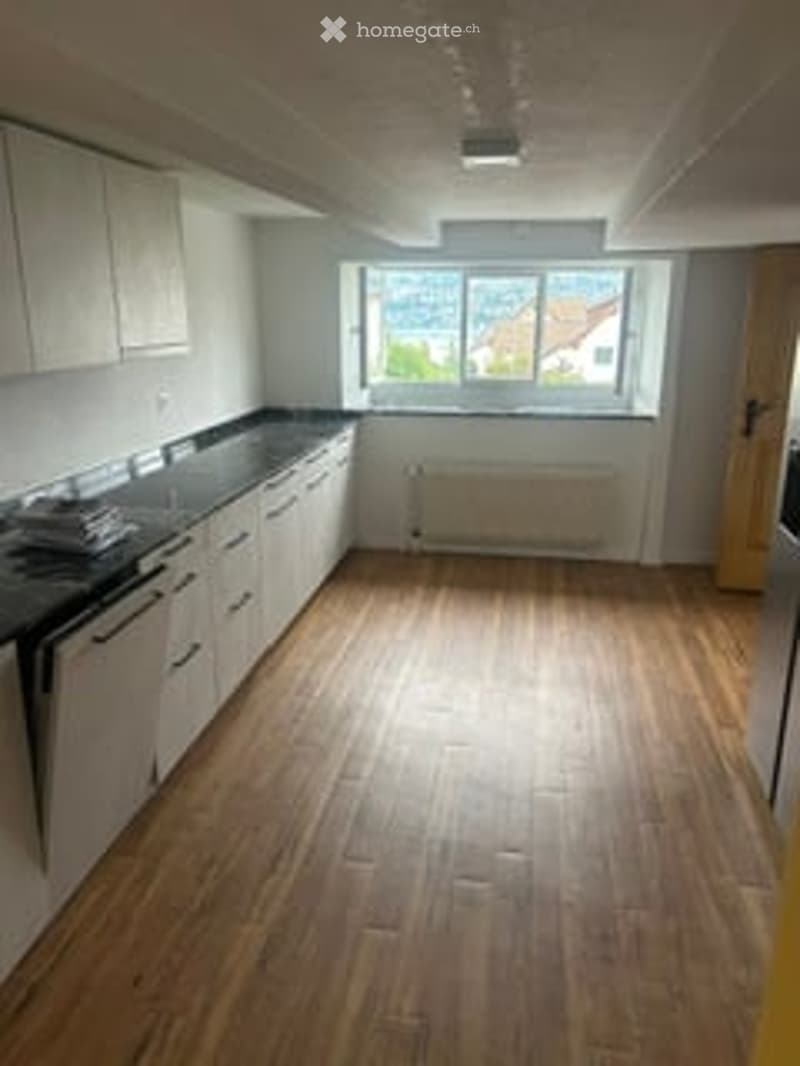 Empty kitchen with white cabinets, black countertop, wooden flooring, and large windows overlooking a view.