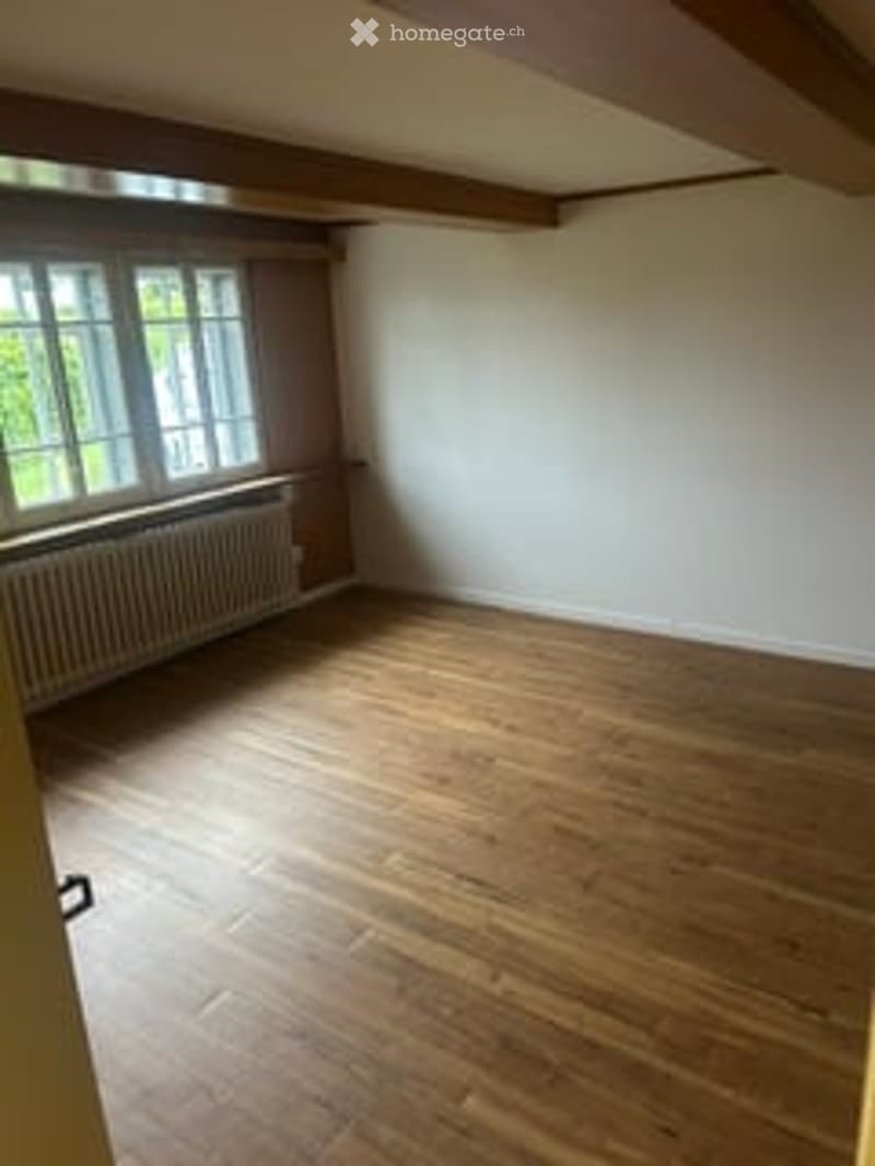 Empty room with wooden flooring, white walls, large window with radiator below, wooden beams on the ceiling.