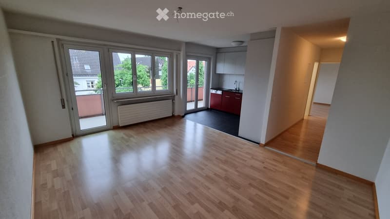 Bright, open-plan living and dining area with wooden flooring, large windows, and a small kitchen area with cabinets and a sink.