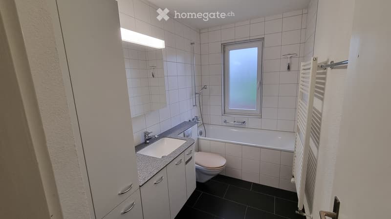 Modern bathroom with white tiles, a bathtub, toilet, sink, mirror, and a radiator. Features a frosted window and a grey countertop.