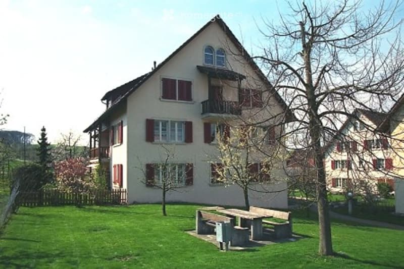 Two-story house with a pitched roof, multiple windows, and a small balcony. The house is surrounded by a well-maintained lawn with a picnic table and benches.