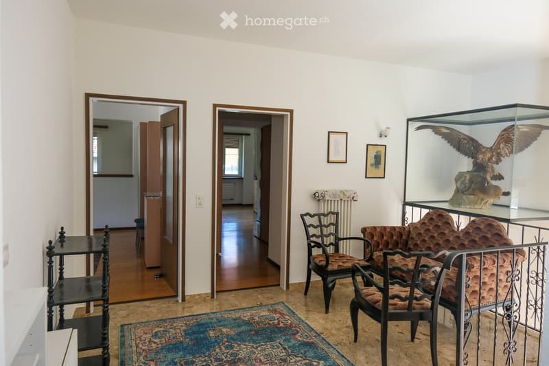 A well-lit room with a patterned rug, a sofa, two chairs, a small table, a display case with a bird sculpture, framed pictures on the wall, and a radiator. The room has wooden flooring and two open doorways leading to other spaces.