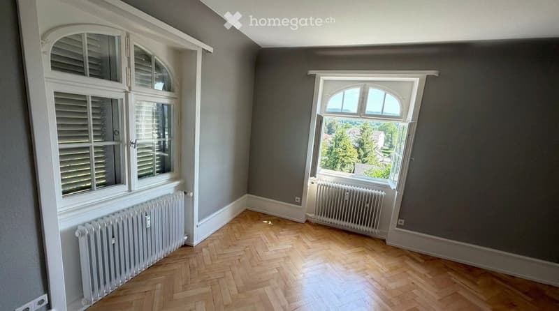 Empty room with wooden herringbone flooring, grey walls, white radiators, and large windows offering a view of greenery outside.