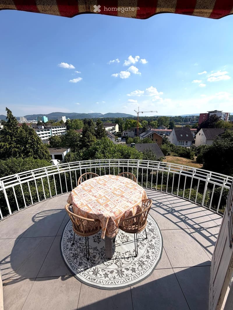 Balcony with a round table and two chairs, overlooking a cityscape with trees and buildings.