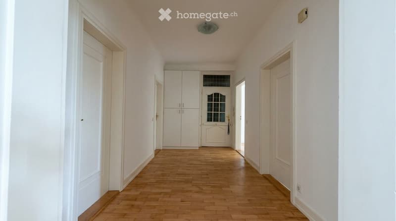 A long, narrow hallway with light wooden flooring, white walls, and several doors leading to different rooms.