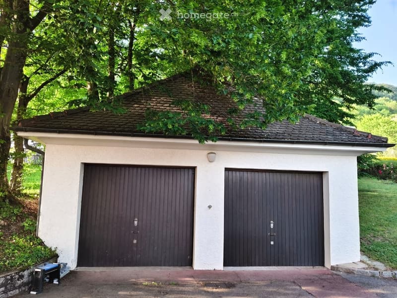A small, white building with two dark wooden garage doors, surrounded by greenery and trees.