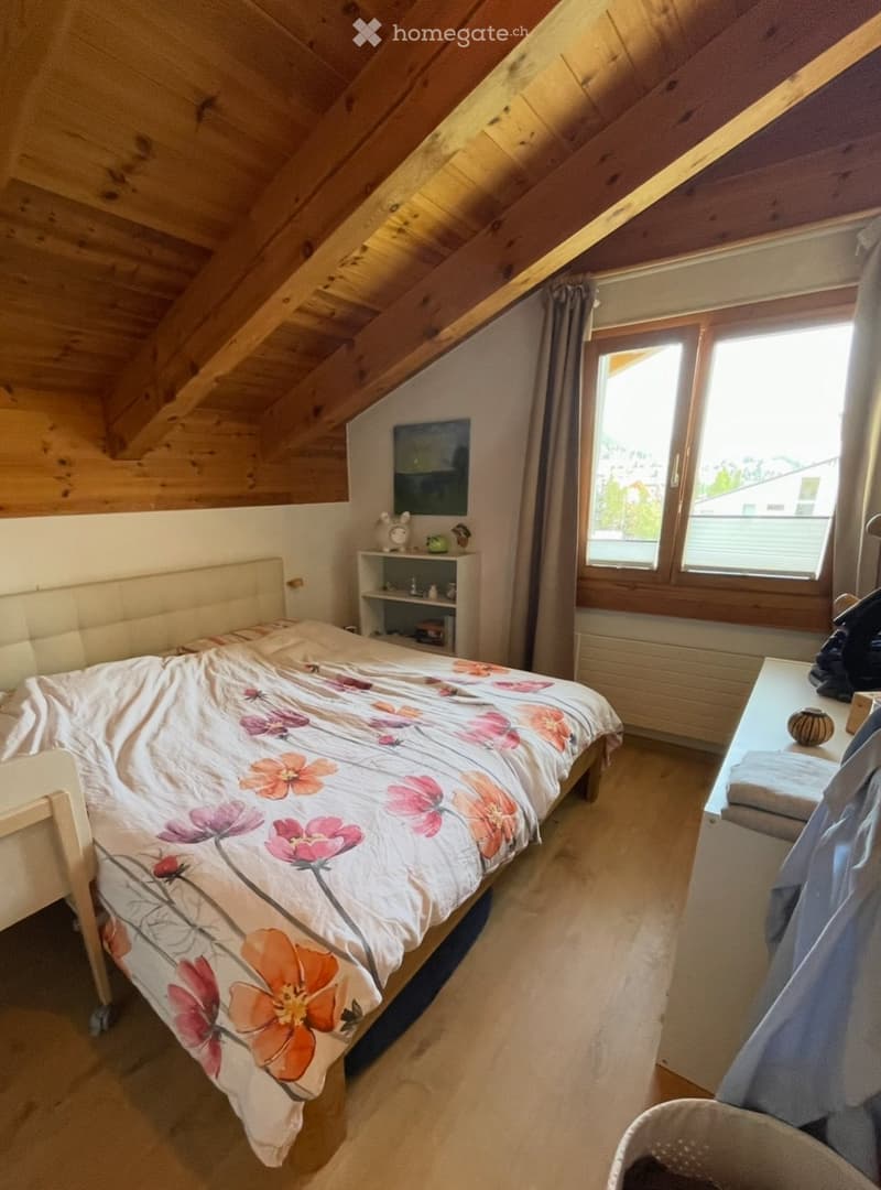 Cozy attic bedroom with wooden slanted ceilings, a large window with curtains, a bed with a floral duvet, a nightstand, a small shelf, and a chair.