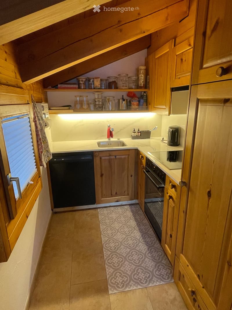 Cozy kitchen with wooden cabinets, a sink, oven, dishwasher, and a small window. Shelves hold jars and containers, and a patterned rug lies on the floor.