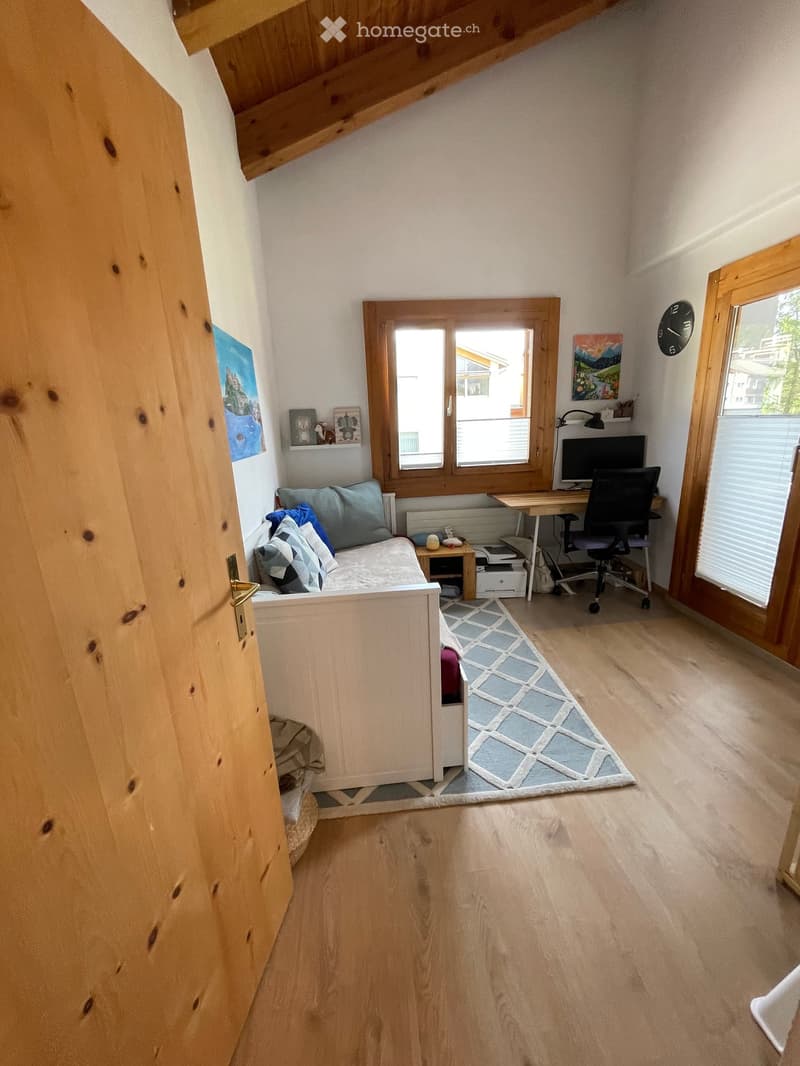 A cozy, multi-functional room with a small sofa, desk, computer, and artwork. The room features wooden floors, large windows, and a modern design.