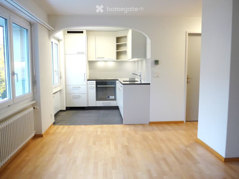 Empty room with light wooden flooring, white walls, and a small kitchen area with white cabinets, a sink, and an oven. The kitchen features a black countertop and a window with blinds.