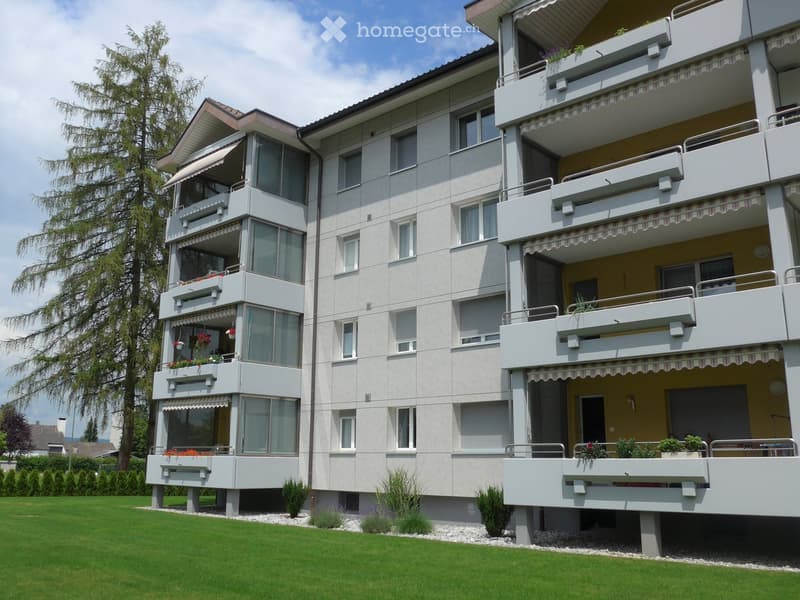 Modern multi-story apartment building with balconies, large windows, and decorative flower boxes. The building has a light-colored facade and is surrounded by a well-maintained lawn and trees.