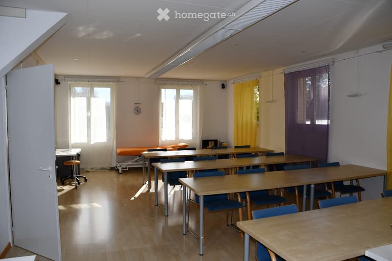 Brightly lit room with wooden flooring, multiple tables and chairs arranged in rows, a small stool, and curtains on the windows.