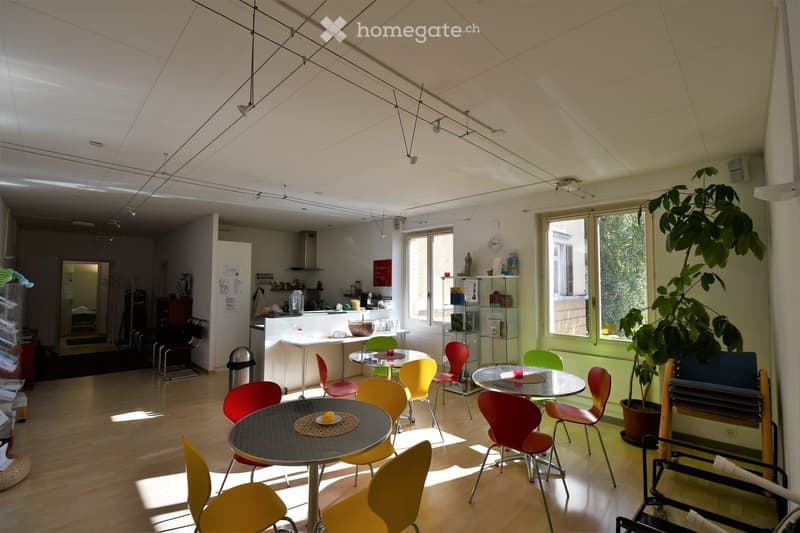 Bright, open-plan room with wooden flooring, large windows, and modern furniture including tables and colorful chairs. A kitchen area is visible in the background with a refrigerator, counter, and various items.