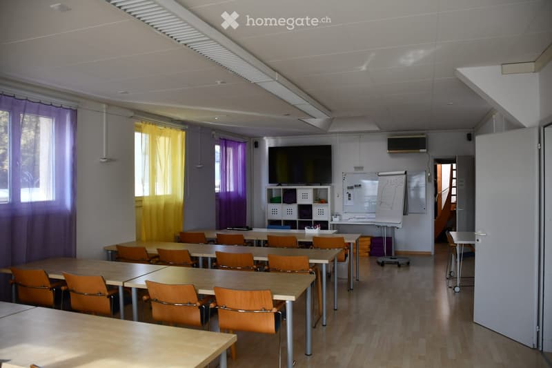 Classroom with multiple tables and orange chairs arranged in rows, a large screen mounted on the wall, a whiteboard, and a small cabinet with storage compartments.
