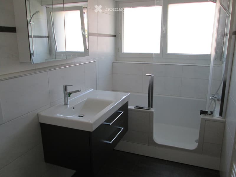 Modern bathroom with white tiles, a white sink with a chrome faucet, a dark wooden cabinet, a bathtub, and a large mirror above the sink.