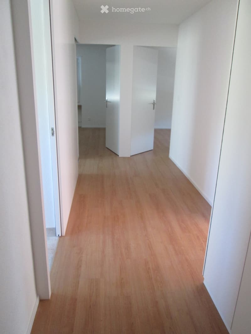 Empty hallway with light wooden flooring and white walls, featuring two closed doors on the right side and an open doorway on the left leading to another room.