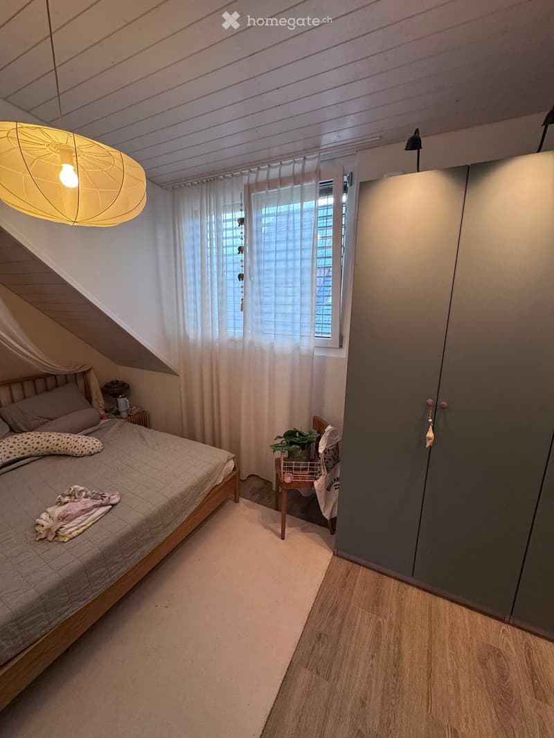 Cozy attic bedroom with wooden slanted ceiling, large window with white curtains, bed with grey bedding, nightstand, hanging lamp, small plant on a chair, and built-in wardrobe.