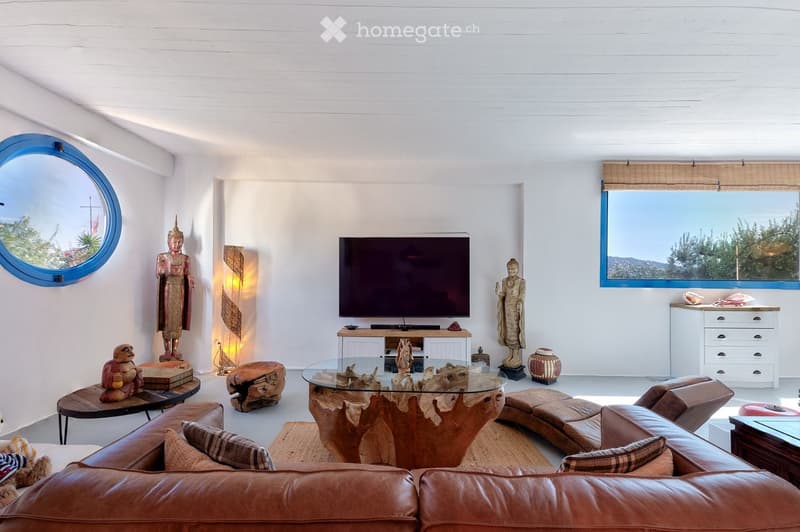 Modern living room with white walls, large windows offering a scenic view, leather sofa, glass coffee table, TV, decorative statues, and a floor lamp.
