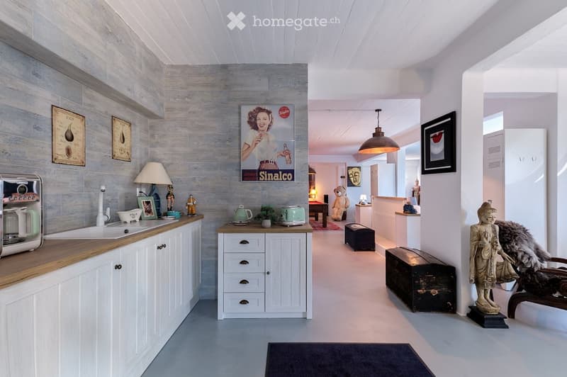 A modern kitchen with white cabinets, wooden countertops, and various appliances. The space features a sink, coffee maker, teapot, toaster, and decorative items including framed pictures, a lamp, and a teddy bear. A small table and chair are visible in th