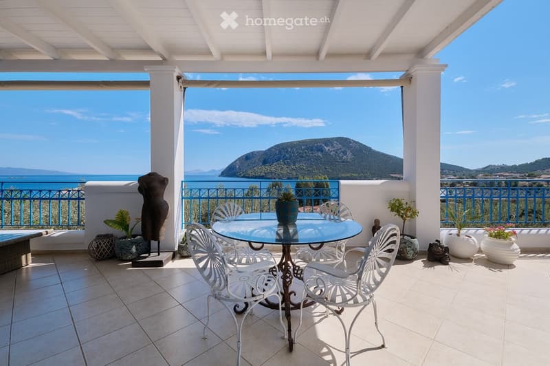 A sunny balcony with a glass table, white metal chairs, potted plants, and a scenic sea and mountain view.