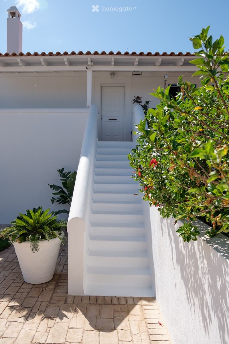 White staircase leading to a door, surrounded by greenery and a large potted plant, with a covered walkway and a small chimney on the roof.