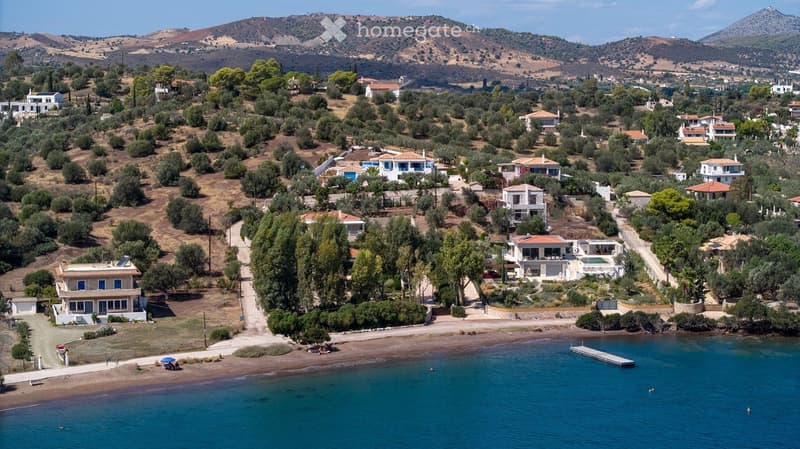 Aerial view of a coastal village with houses, trees, and a beach, surrounded by hills and mountains.