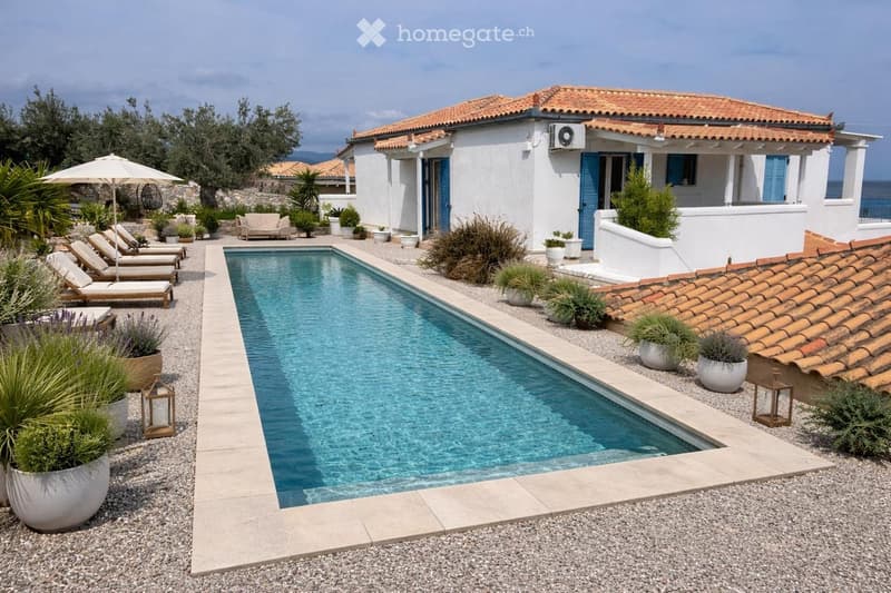 A modern outdoor area with a rectangular swimming pool, surrounded by potted plants, lounge chairs, and a white umbrella. The area is bordered by gravel and features a white house with blue shutters in the background.